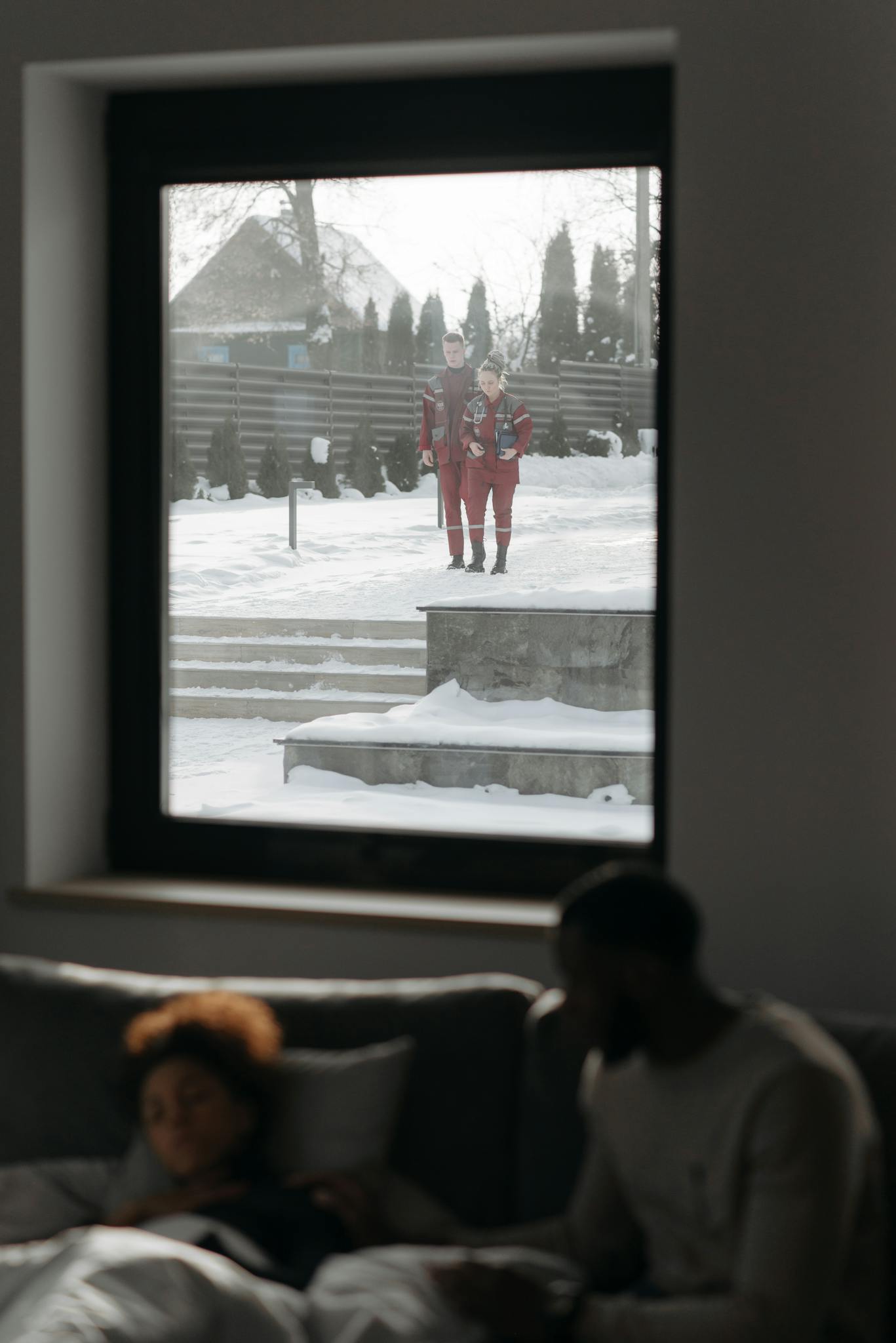 Paramedics view through a window as they approach a home during winter. Indoors, a patient rests.